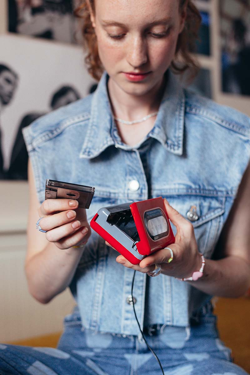 A girl with red hair in a jean vest puts a cassette tape into a Walkman to listen to.