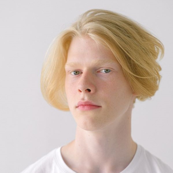 A teenage boy in a white shirt with pale blonde hair parted down the center, grown out to the ears and swept back looks at the camera and slightly smiles.