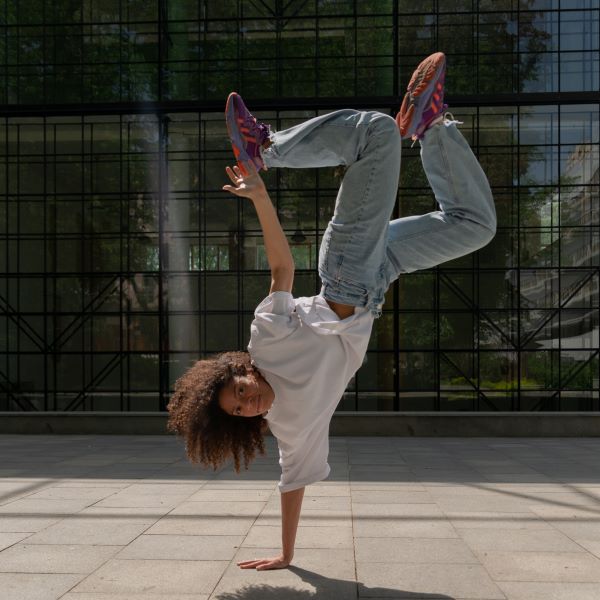 A woman in a baggy white tee shirt, light denim pants, and multicolored sneakers does a one-handed handstand on the sidewalk.