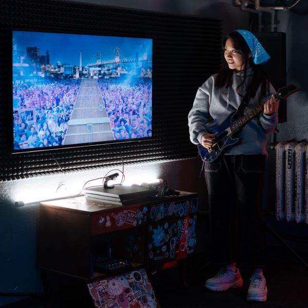 A woman in a blue bandana, grey sweatshirt, and white sneakers stands next to a TV while she plays Guitar Hero.