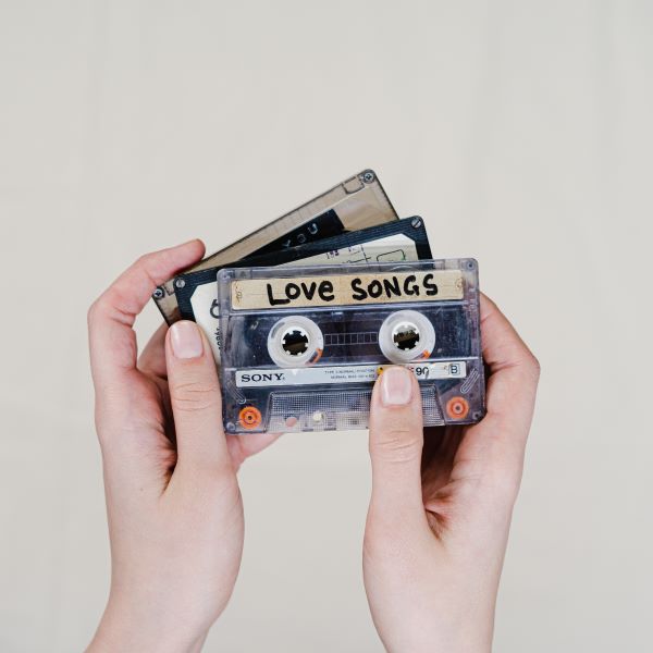 Two hands hold three cassette tapes, with 'Love Songs' written on one in black marker.