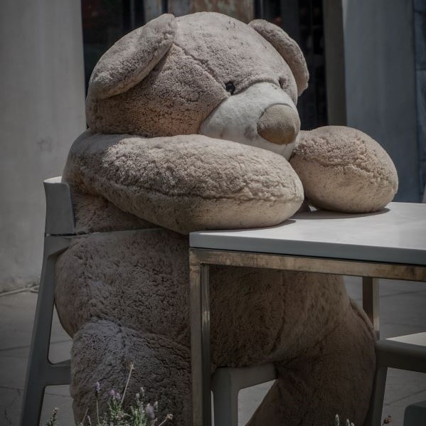 A man-sized teddy bear sits on a chair at a table.